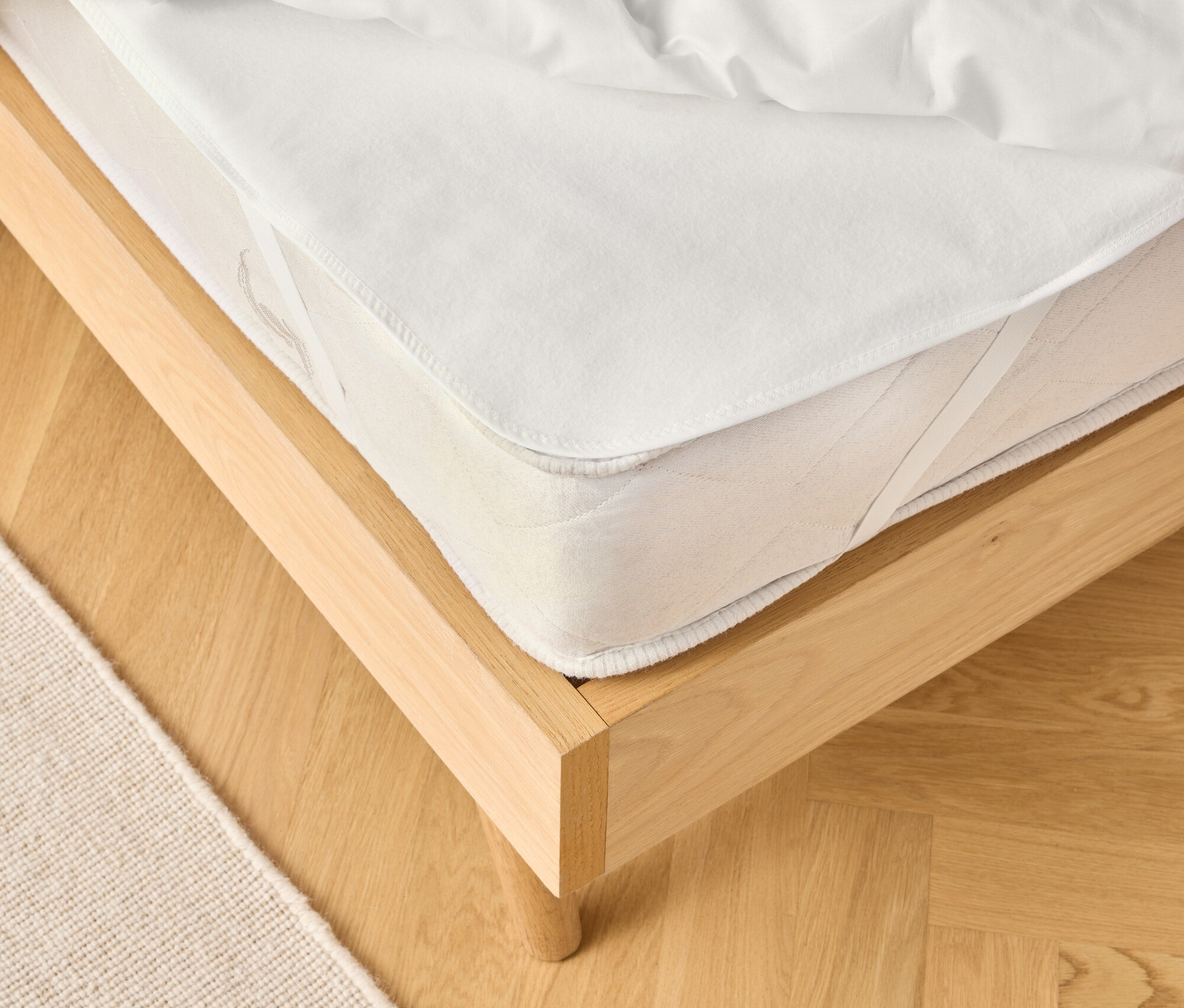 Pohled na světlý dřevěný rám postele s bílou matrací a bílou přikrývkou. Béžový koberec je částečně viditelný.