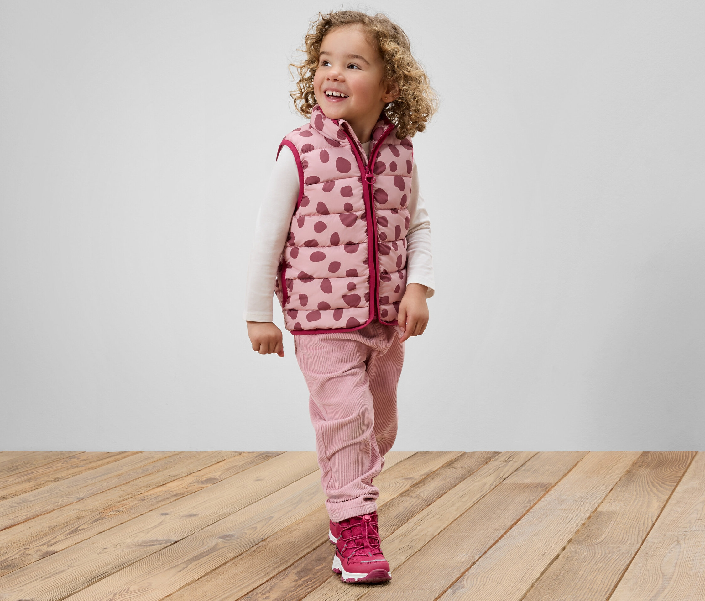Usmívající se holčička s kudrnatými vlasy v růžové vestě s puntíky, růžových kalhotách a růžových botách stojí na dřevěné podlaze.