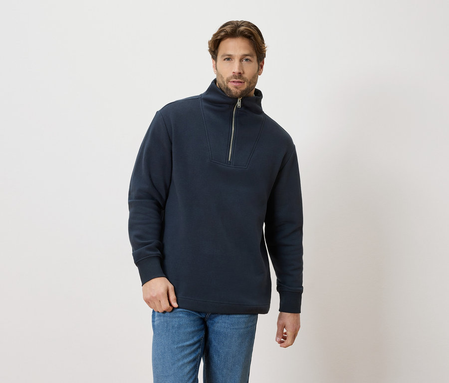 Muž má na sobě tmavě modrý svetr na zip a modré džíny.