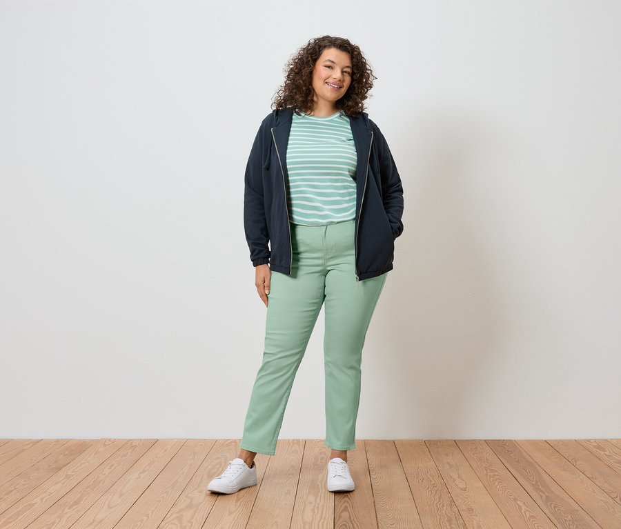 Žena s kudrnatými vlasy stojí před zdí a má na sobě tmavě modrou bundu, pruhované tričko, zelené kalhoty a bílé boty.