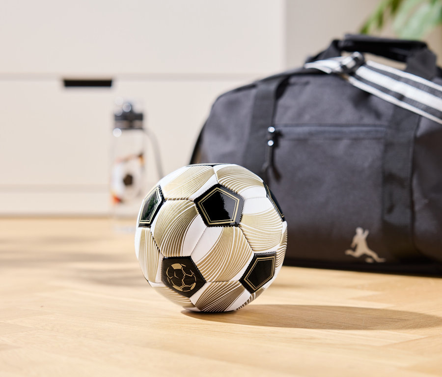 Zlatý fotbalový míč leží na dřevěné podlaze, v pozadí je sportovní taška a láhev na vodu.
