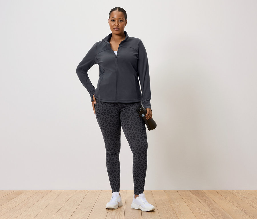 Žena v tmavé sportovní bundě a legínách s leopardím vzorem, drží láhev s vodou.