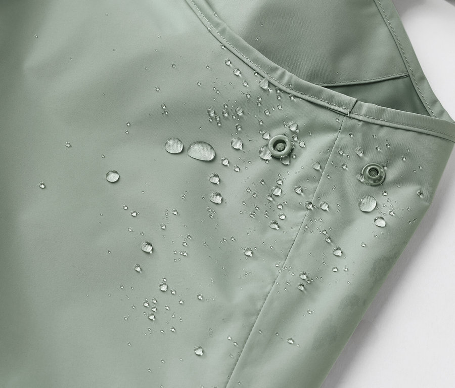 Detailní záběr na voděodolnou, světle zelenou látku s kapkami vody a dvěma patentkami.