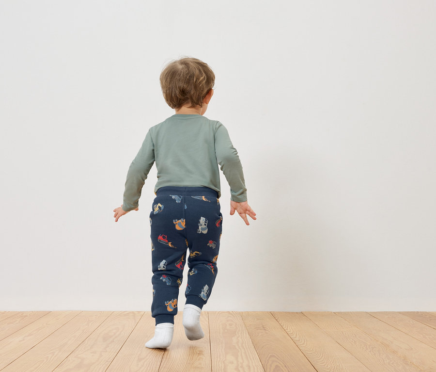 Malé dítě s blond vlasy, viděné zezadu, má na sobě zelené tričko s dlouhým rukávem a tmavě modré kalhoty se vzory vozidel.