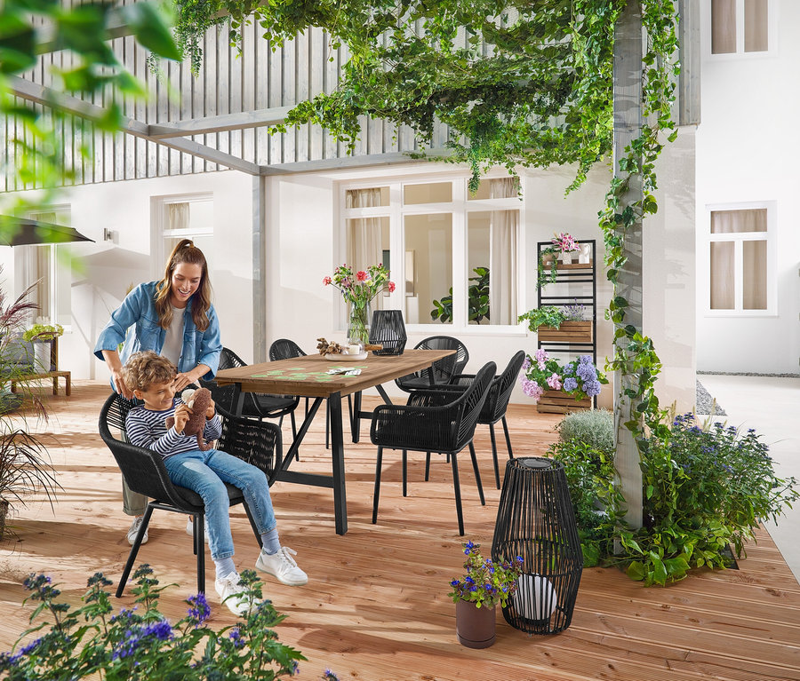 Matka a dítě sedí u Rozkládacího zahradního stolu s rustikální konstrukcí, cca 200–250 cm. Malá a velká Solární lucerna s textilním výpletem stojí na zemi. V pozadí je Zelená stěna s flexibilně zavěsitelnými policemi »Elin«.