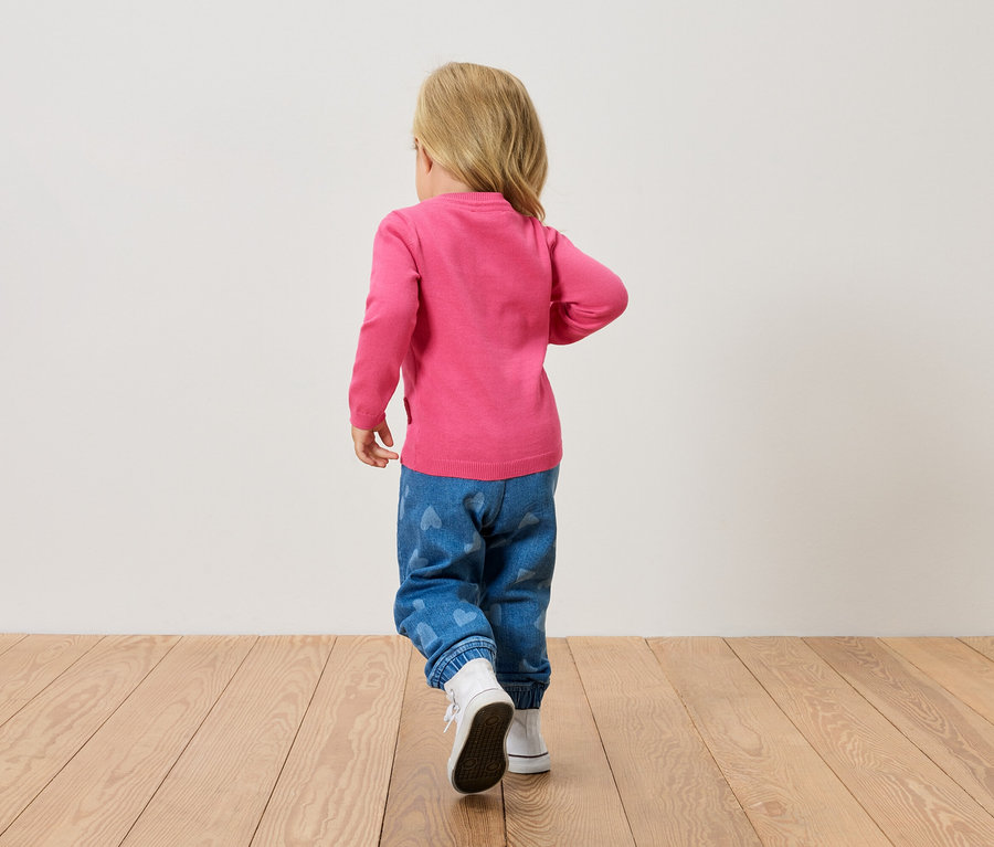 Malé blond dítě zezadu, má na sobě růžový svetr, modré džíny se srdíčky a bílé boty, kráčí po dřevěné podlaze.