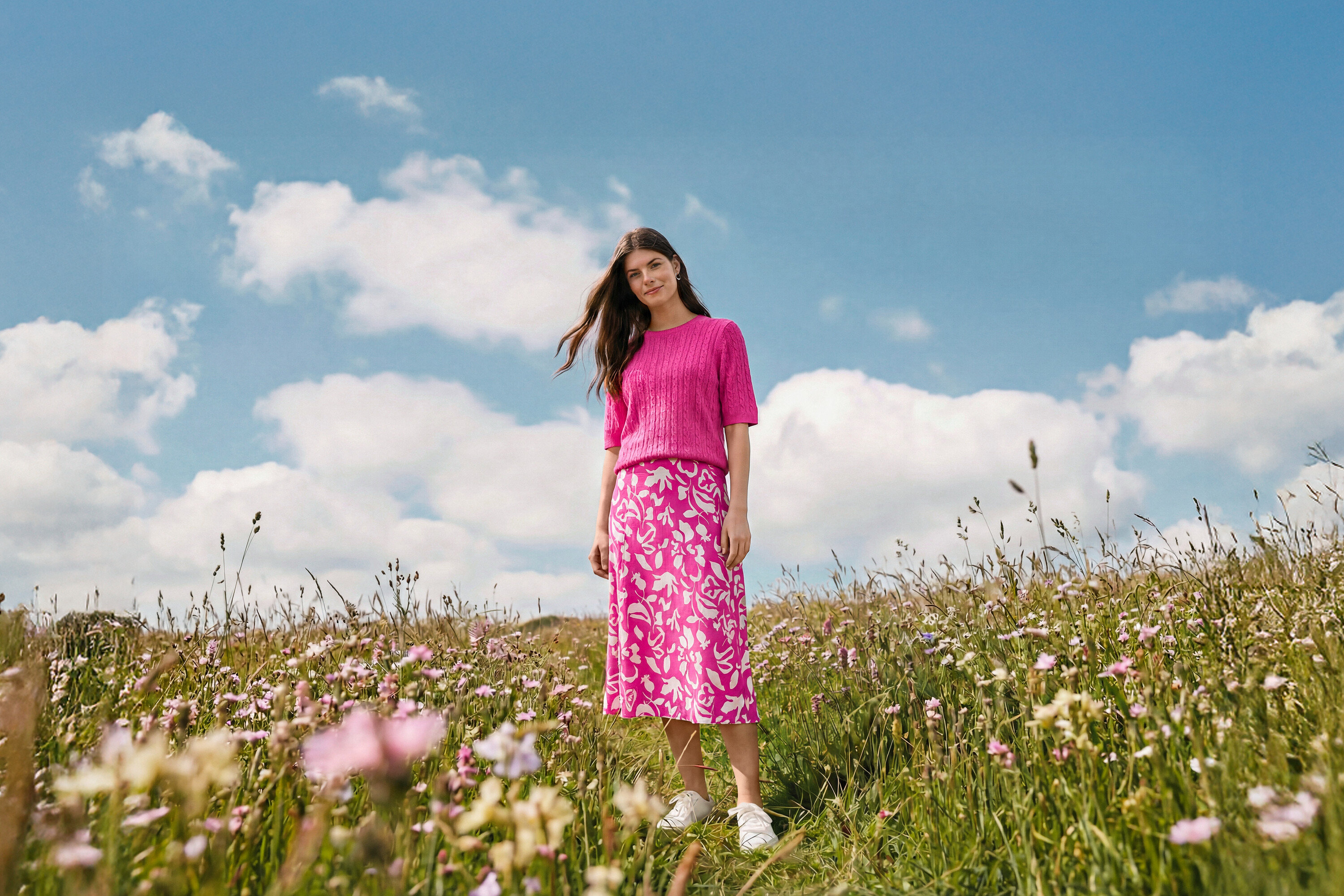 Žena stojí na louce a má na sobě midi sukni s potiskem, náušnice Wave a kožené tenisky.