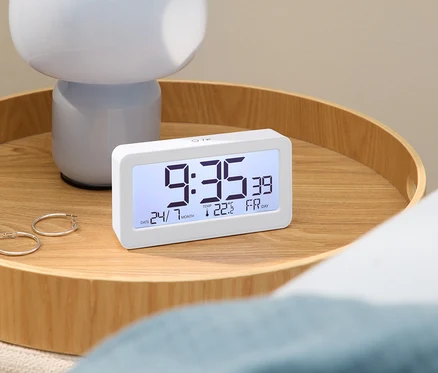 Digitální budík s časem 9:35 na dřevěném stole s lampou a náušnicemi.