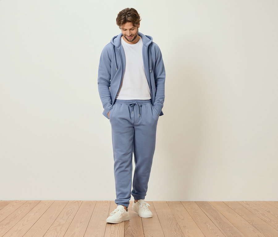 Muž stojí s rukama v kapsách ve světle modré teplákové soupravě a bílých teniskách.