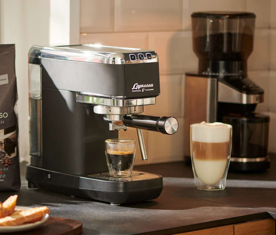 Espresso vytéká z pákového kávovaru »Lapressa«, černý, do sklenice. V pozadí je mlýnek na kávu a balíček kávy.