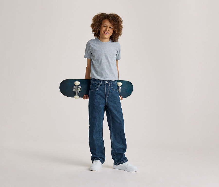 Usmívající se chlapec s kudrnatými vlasy v šedém tričku, modrých džínech a bílých teniskách, držící za zády tmavě modrý skateboard.