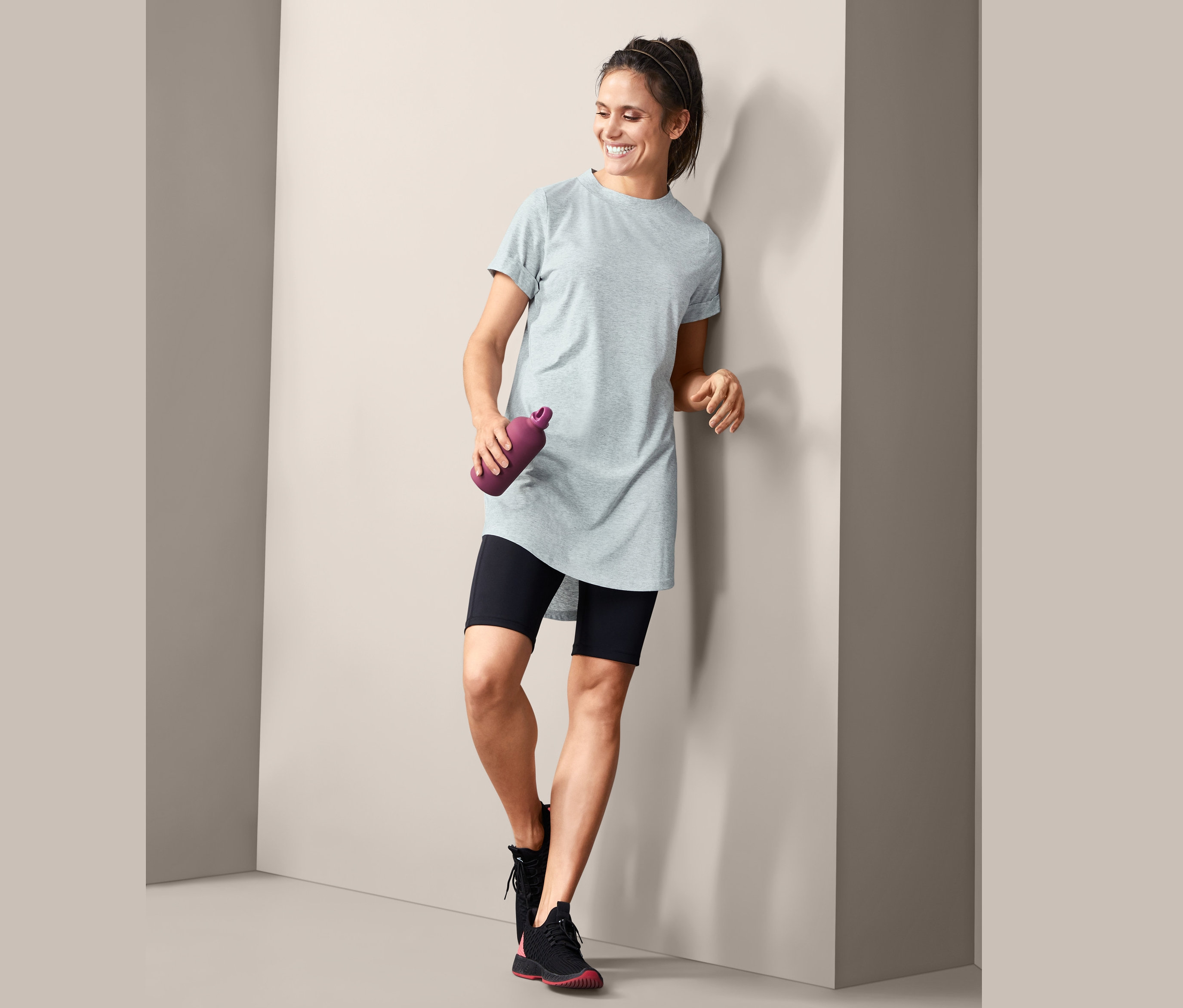 Usmívající se žena opřená o zeď drží fialovou láhev na vodu a má na sobě šedé dlouhé triko.