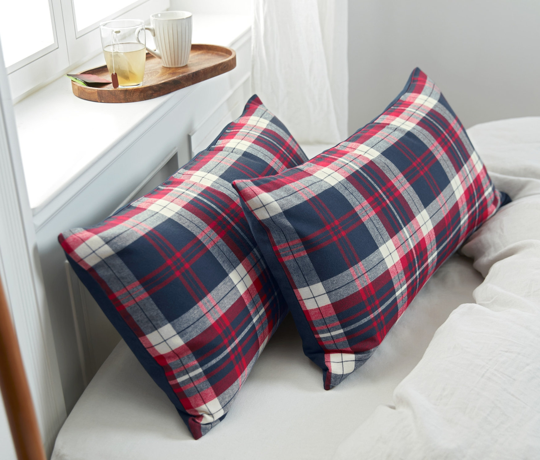 Dva flanelové povlaky na polštáře s červeným, bílým a modrým kostkovaným vzorem leží na posteli. Vedle nich je šedá deka a tác s šálky.