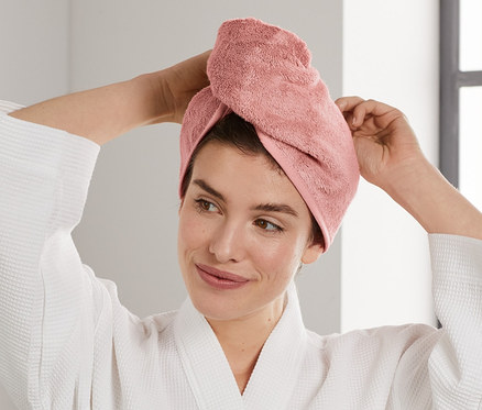 Žena si upravuje růžový ručníkový turban na hlavě. Oblečená v bílém županu stojí v koupelně.