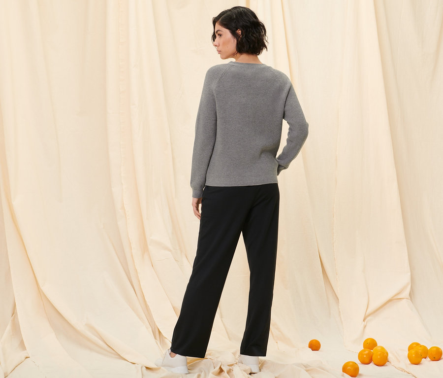 Žena stojí zády k fotoaparátu a má na sobě kardigan z hrubé pleteniny a černé teplákové kalhoty.