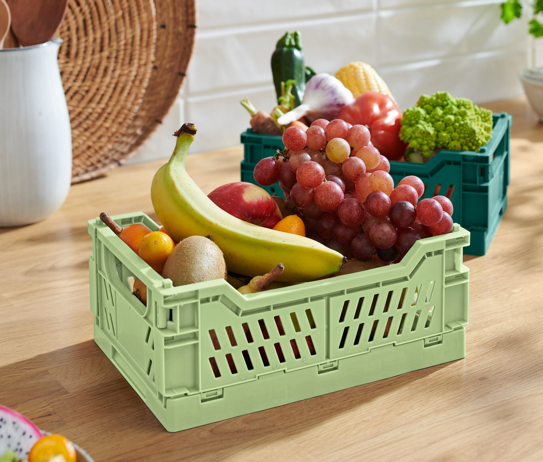 Zelená skládací úložná bedna s banánem, jablkem, kiwi a hrozny. V pozadí je zelená skládací úložná bedna se zeleninou.