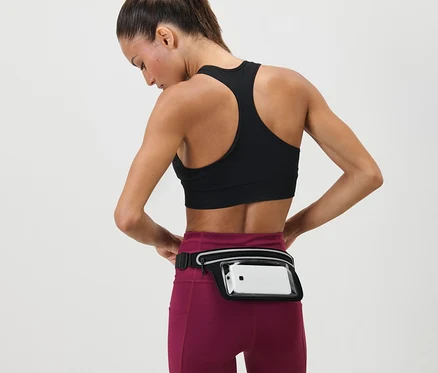 Žena si upravuje Běžecký opasek na smartphone kolem pasu. Je oblečená ve sportovní podprsence a legínách.