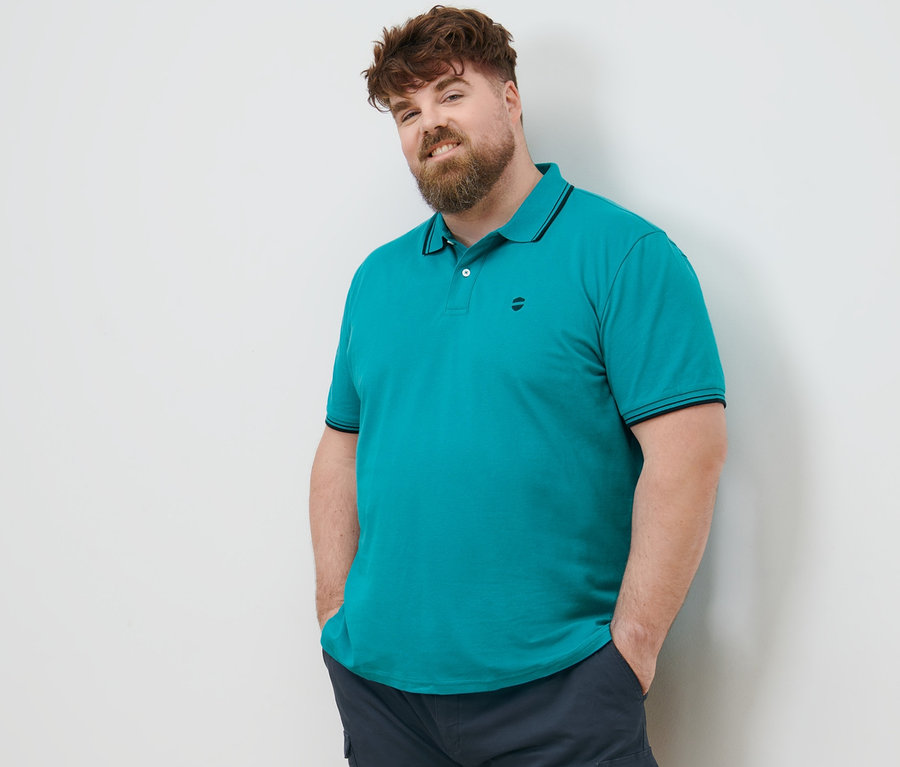 Muž s hnědými vlasy stojí a má ruce v kapsách cargo šortek námořnické modré barvy. Má na sobě modrozelené polo tričko z piké.