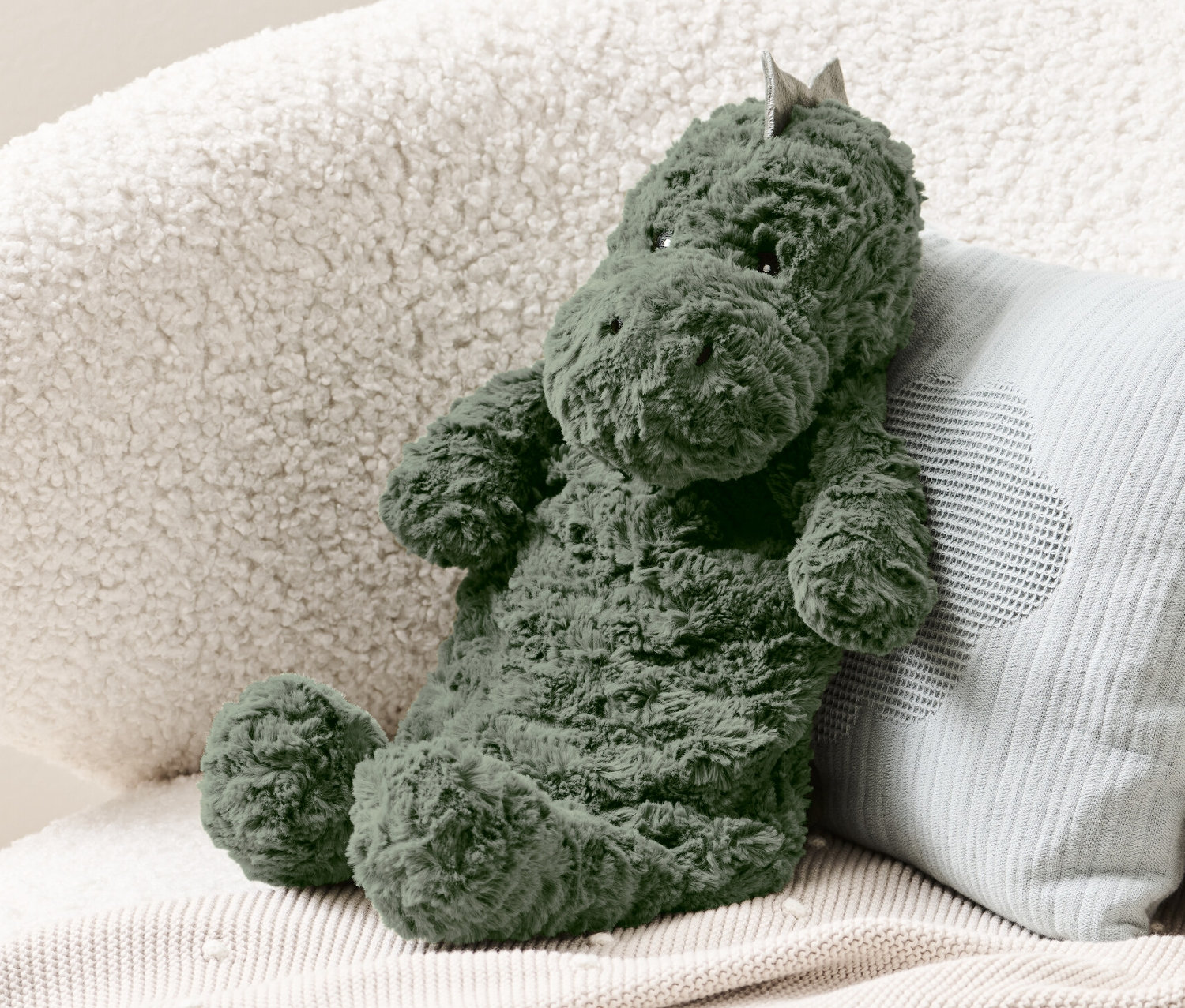 Zelený dinosauří termofor sedí na krémovém křesle vedle světle modrého polštáře.