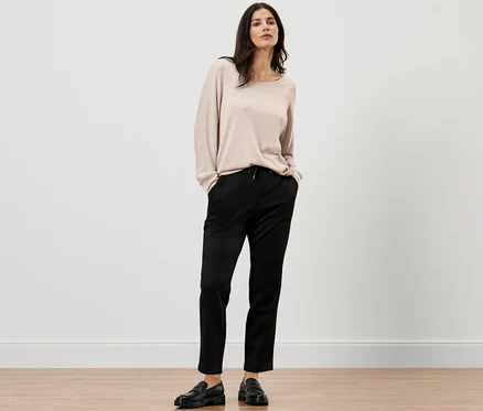 Žena pózuje v béžovém svetru a černých joggpant kalhotách.