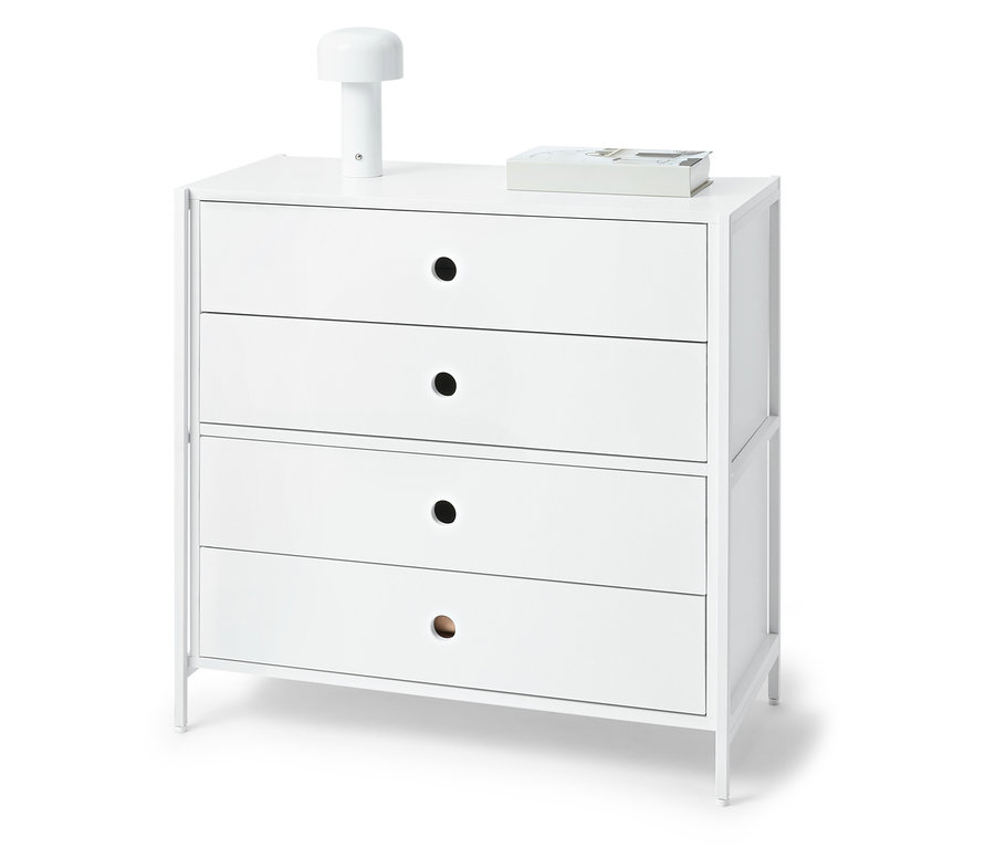 Bílá komoda se čtyřmi zásuvkami, na horní desce lampa a kniha.