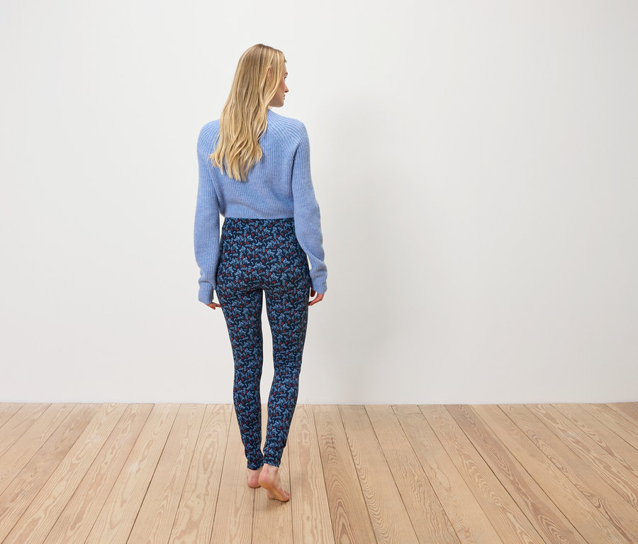 Žena v modrém svetru a vzorovaných legínách stojí zády k fotoaparátu.