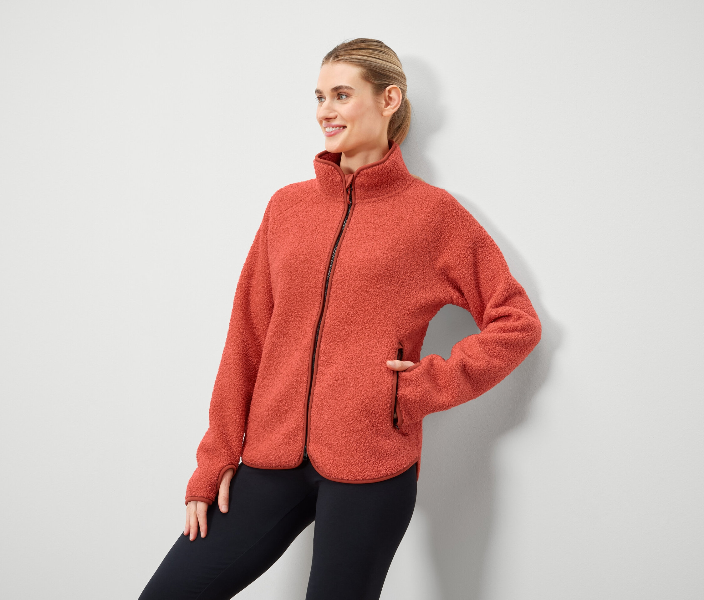 Žena pózuje v oranžové fleecové bundě na zip a černých legínách.