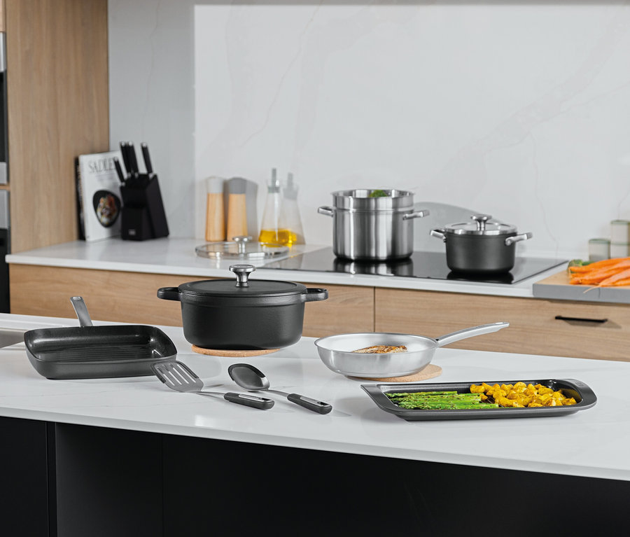 Různé hrnce, pánve a kuchyňské náčiní na bílé kuchyňské desce.