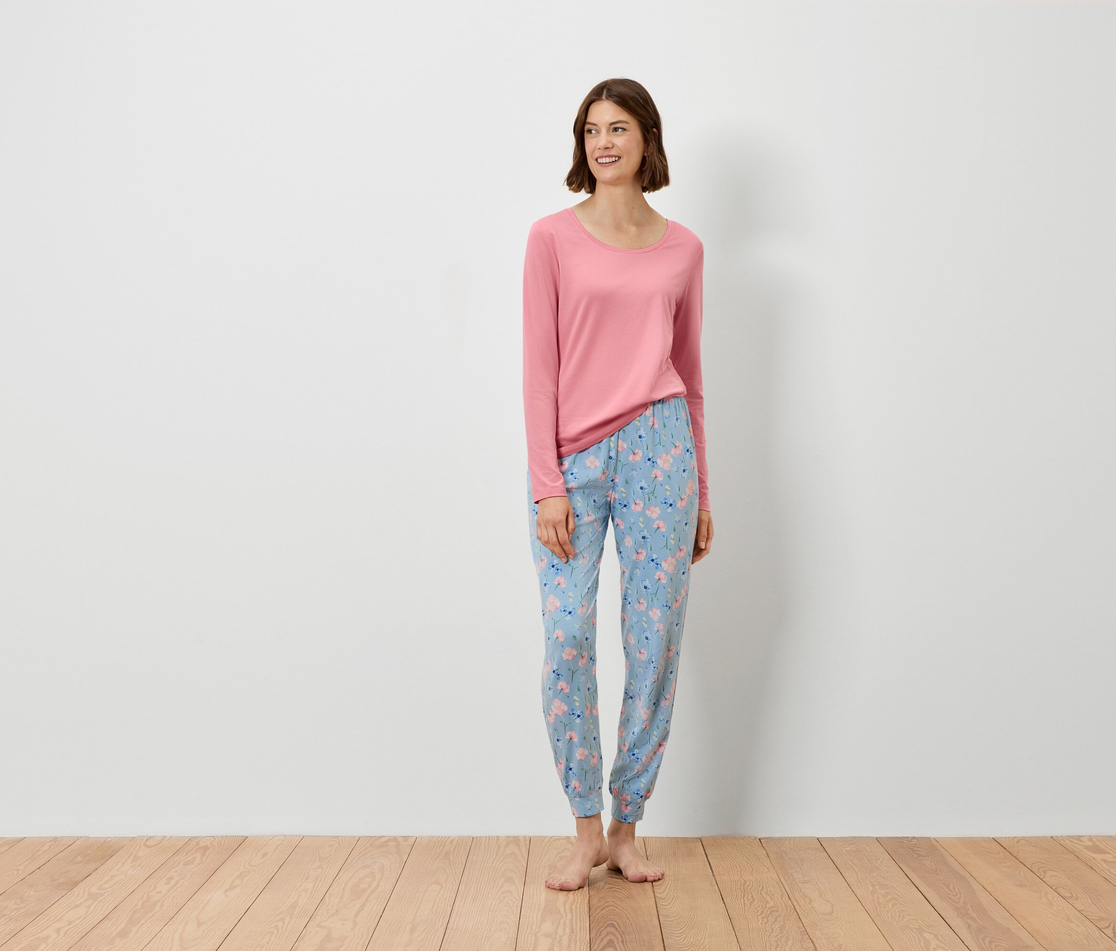 Žena stojí a má na sobě růžový top s dlouhým rukávem a modré kalhoty s květinovým vzorem.