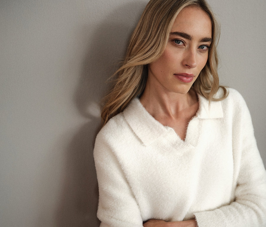 Žena s blond vlasy v bílém pleteném svetru s polo límečkem Street One se opírá o šedou zeď.