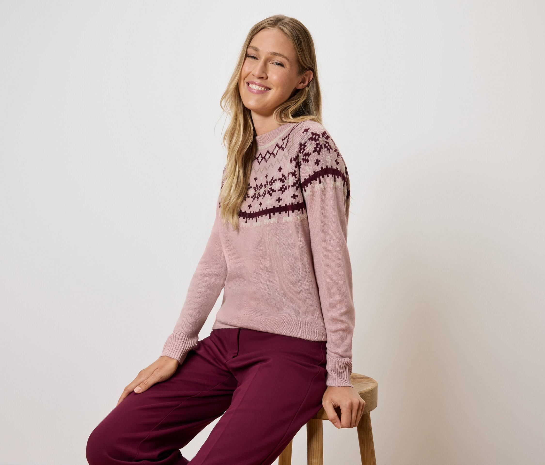 Žena sedí na stoličce a má na sobě růžový svetr se vzorem a bordó kalhoty.