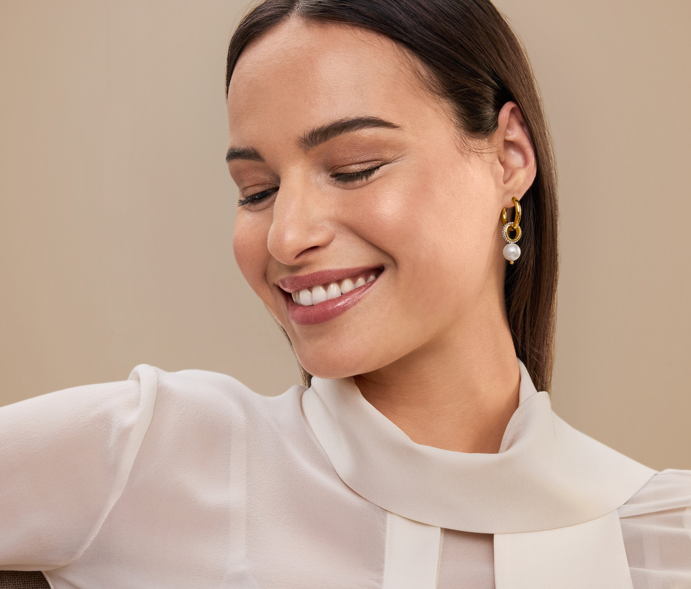 Usmívající se žena s hnědými vlasy a zlatými náušnicemi s perlou, má na sobě bílou blůzu.