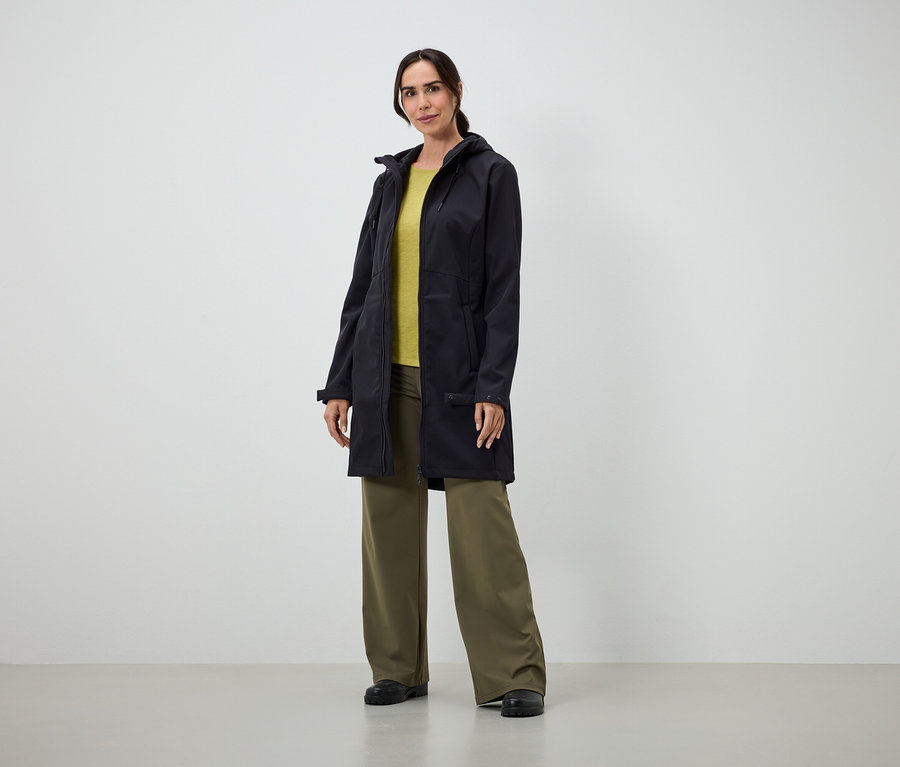 Žena v dlouhém černém kabátu, žlutém tričku a širokých zelených kalhotách stojí v interiéru.