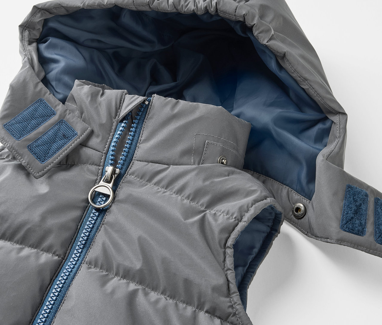 Detail šedé prošívané vesty s kapucí Reflexní dětská prošívaná vesta má modrý zip a modrou vnitřní stranu kapuce.