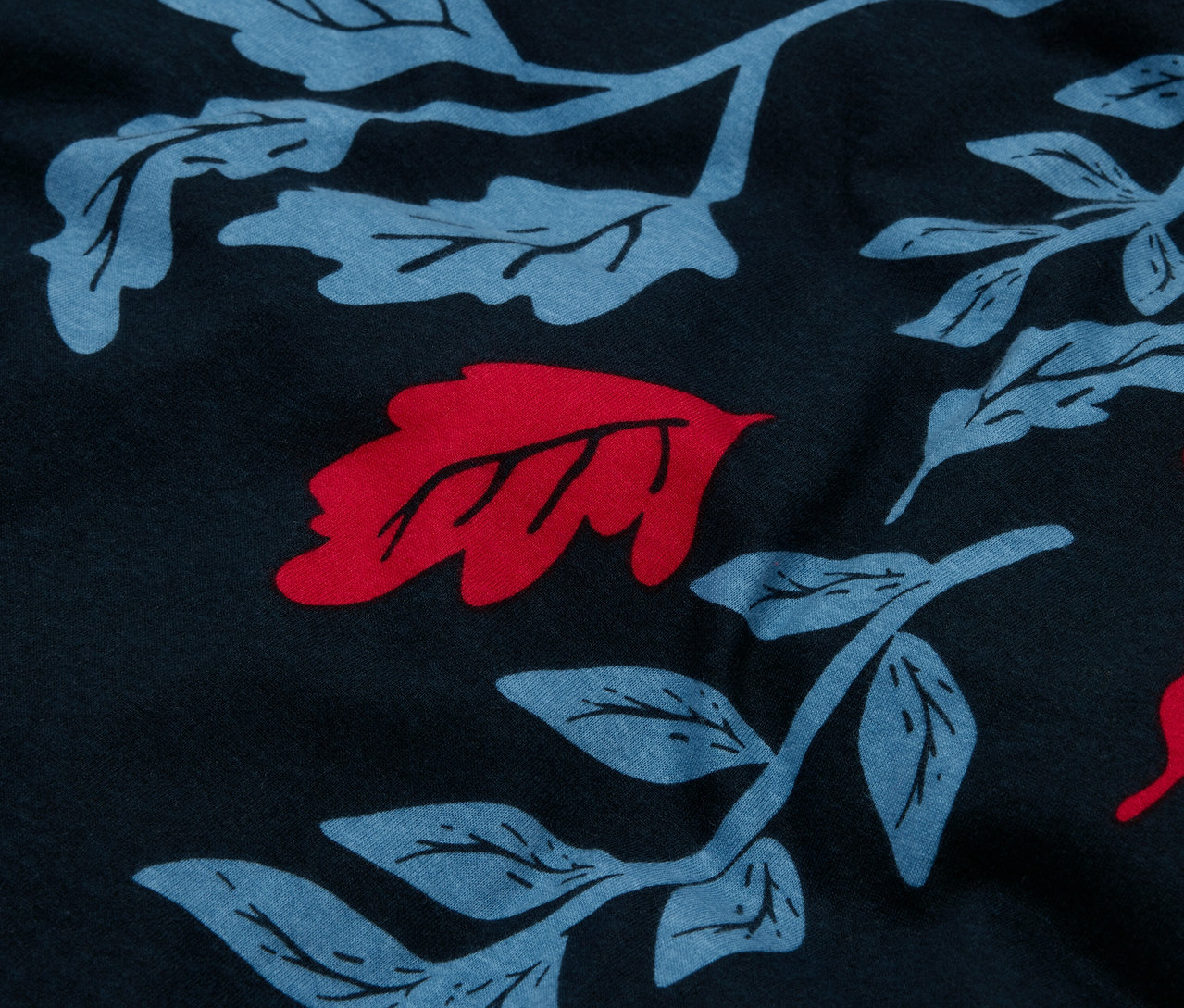 Detailní záběr na tmavě modrou látku s celoplošným potiskem modrých listů a červených akcentů.