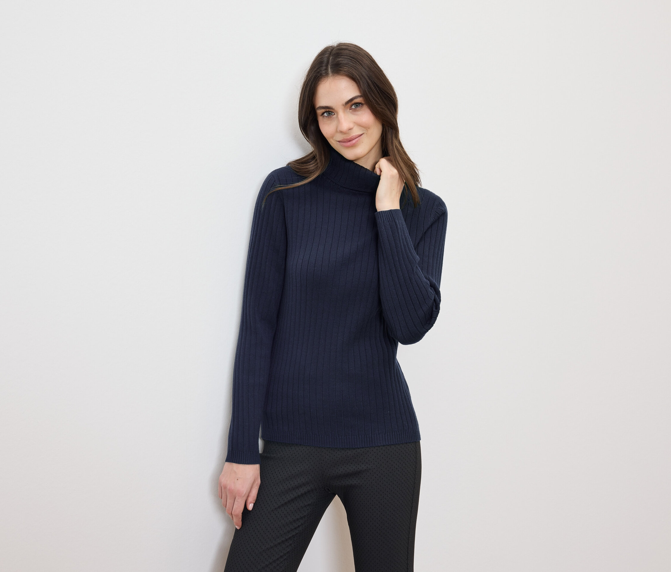 Žena s hnědými vlasy pózuje v tmavě modrém roláku a černých kalhotách.