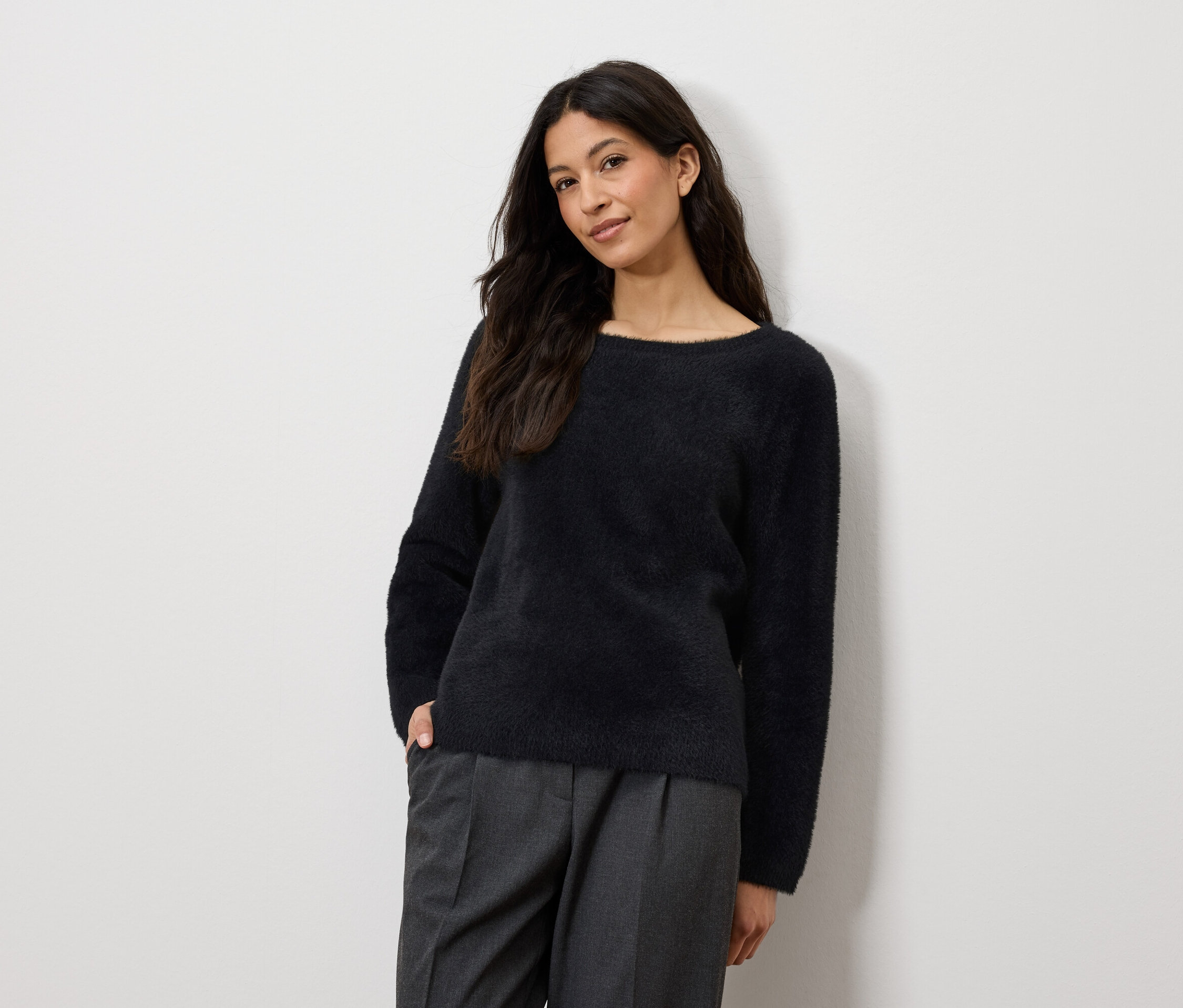 Žena pózuje v tmavě modrém svetru a šedých kalhotách.