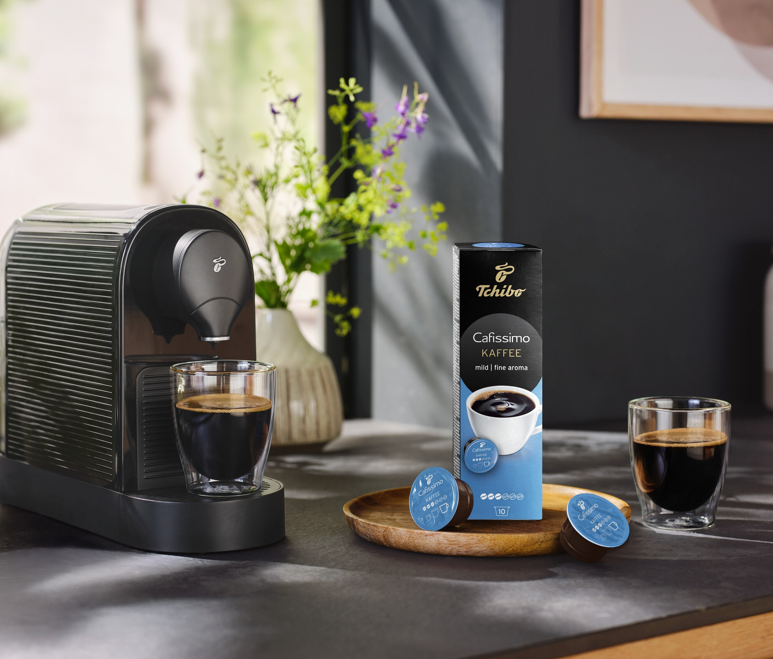 Kávovar stojí vedle sklenice kávy a balení 10 kapslí jemné kávy.