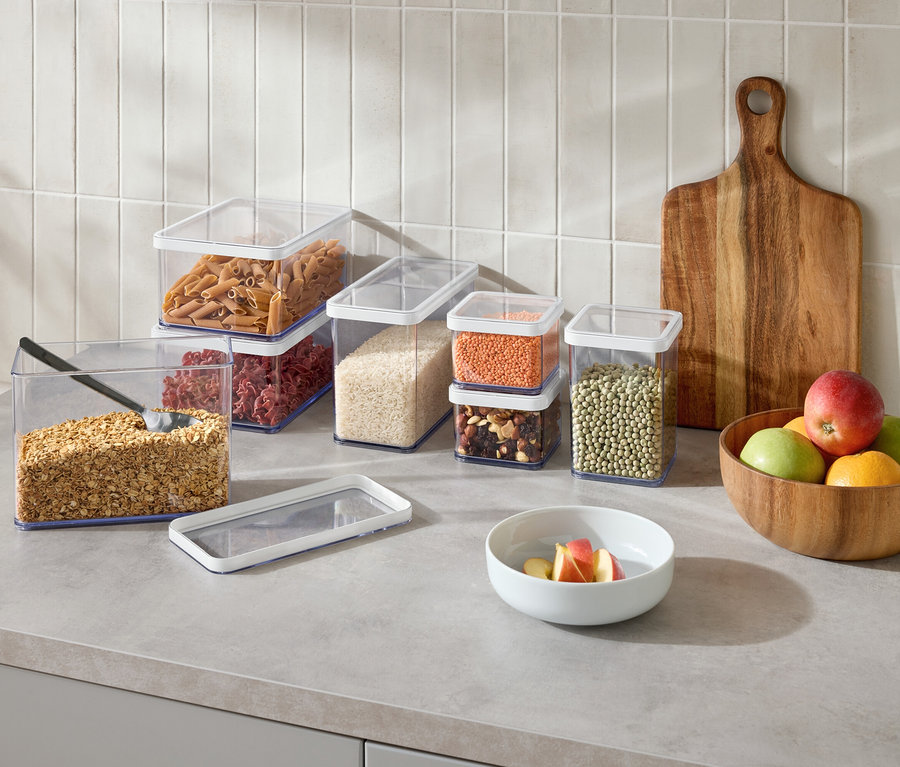 Na kuchyňské lince stojí několik průhledných nádob na potraviny s různými potravinami, dřevěné prkénko a mísa s ovocem.