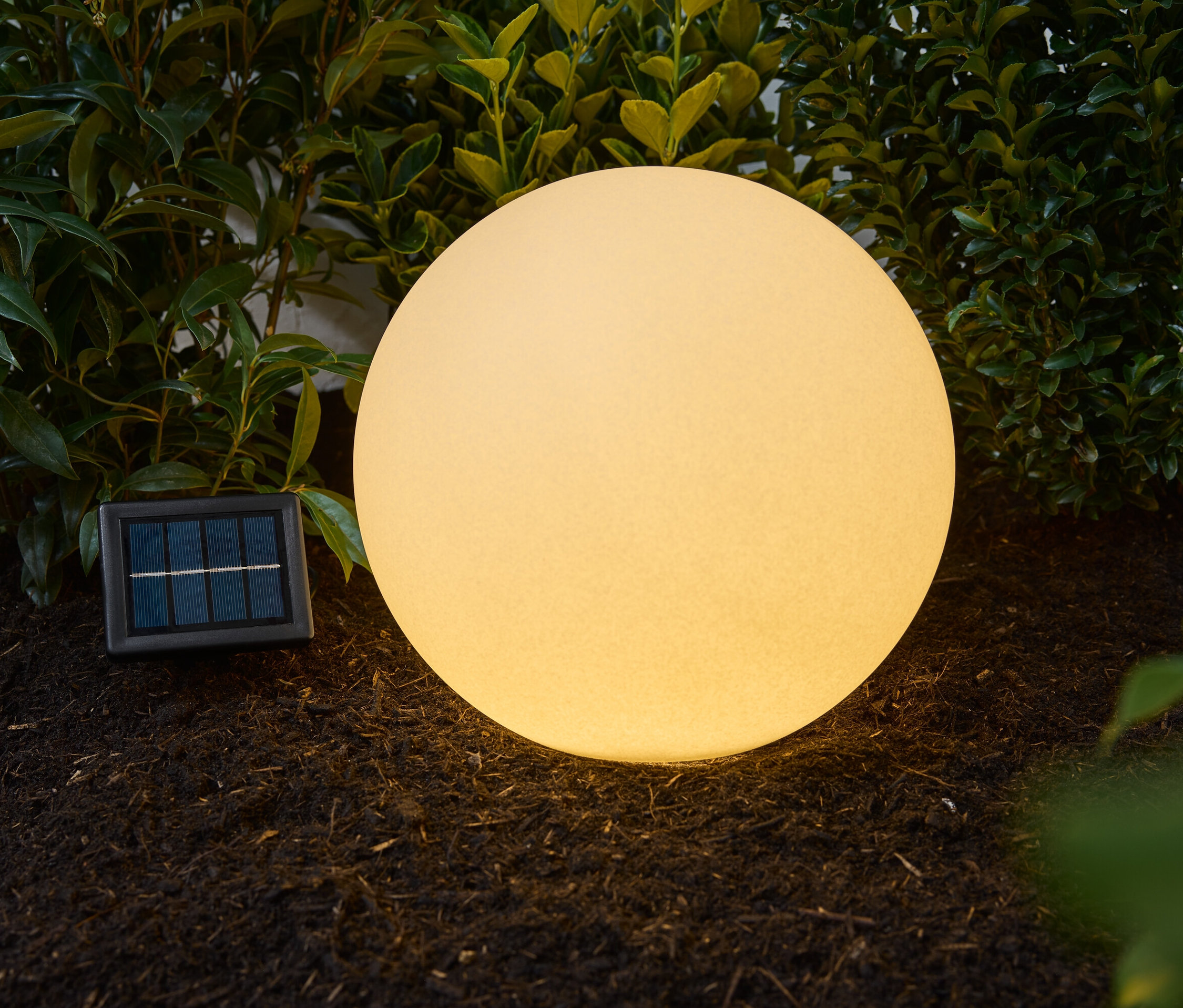 Solární svítidlo ve tvaru koule s pískovým vzhledem, o průměru cca 30 cm, umístěné venku na zemi vedle zelených rostlin.