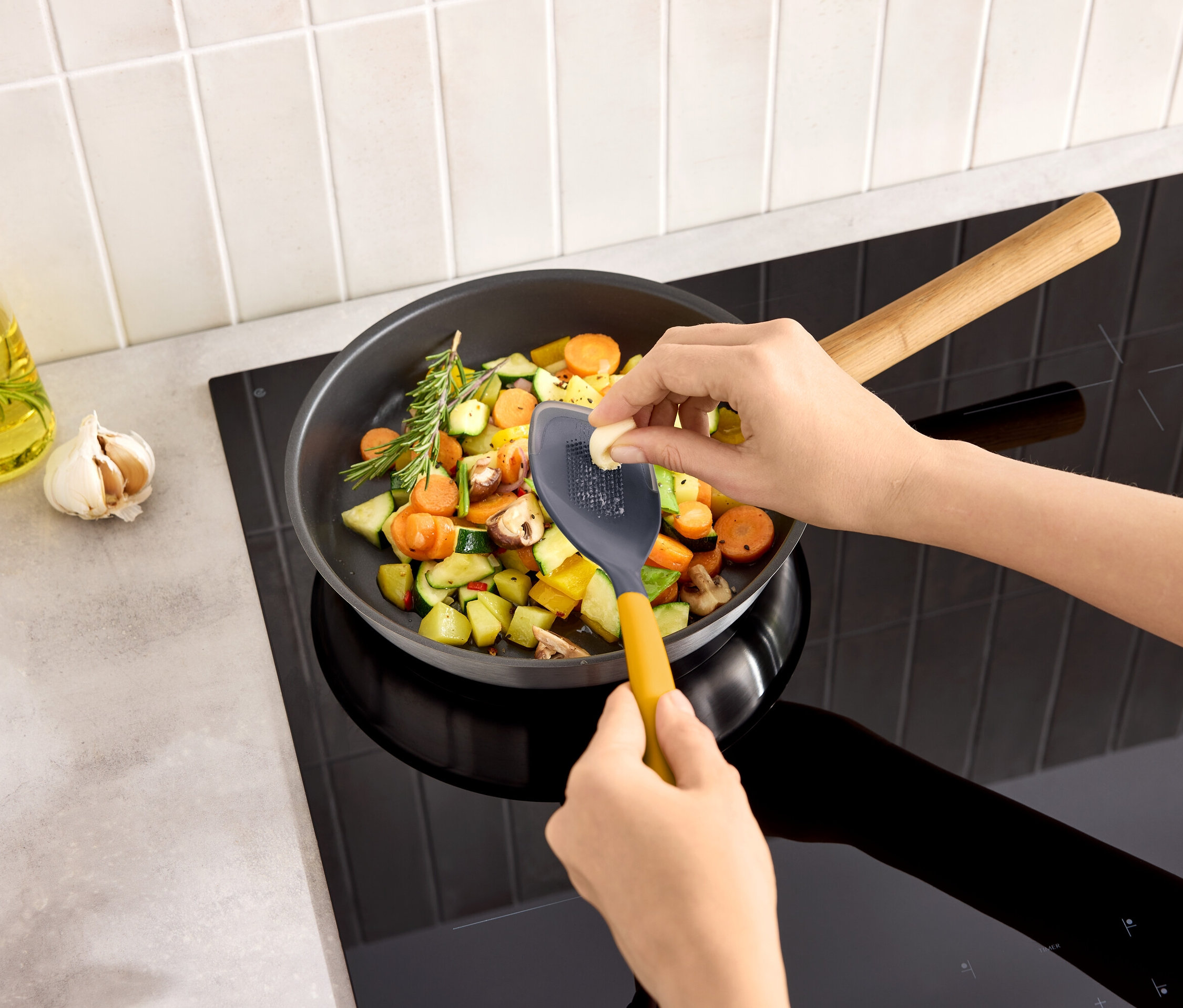 Osoba strouhá stroužek česneku do pánve se zeleninou na varné desce.