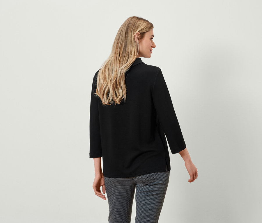 Zezadu blond žena v černém topu s tříčtvrtečními rukávy a šedých kostkovaných kalhotách.