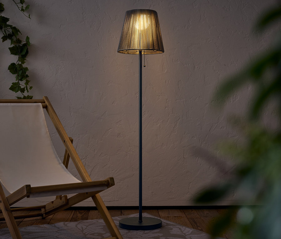 Osvětlená stojací lampa se stínidlem stojí vedle dřevěného lehátka.