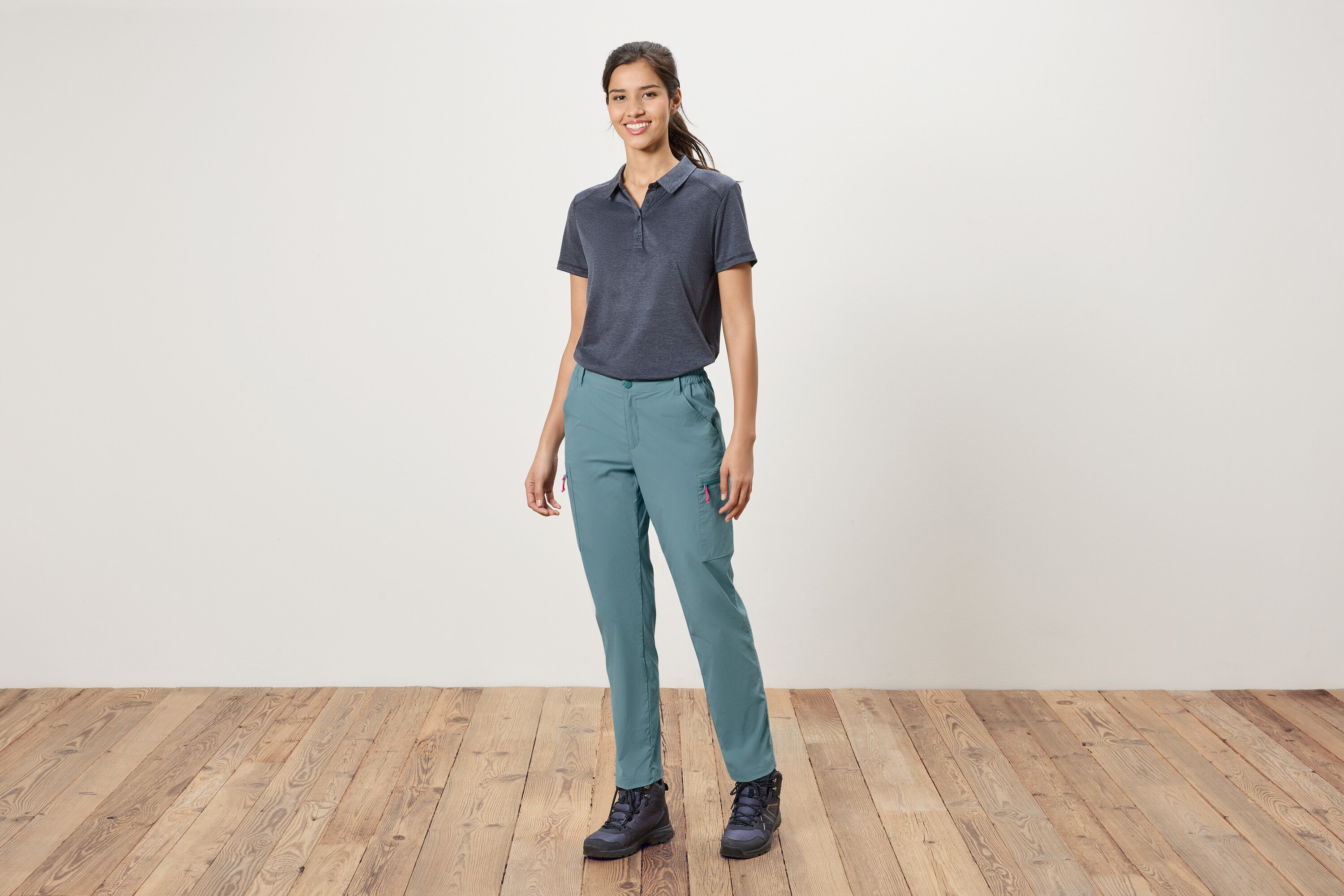 Žena stojí před bílou zdí a má na sobě modré polokošile, modré kalhoty a trekové boty.