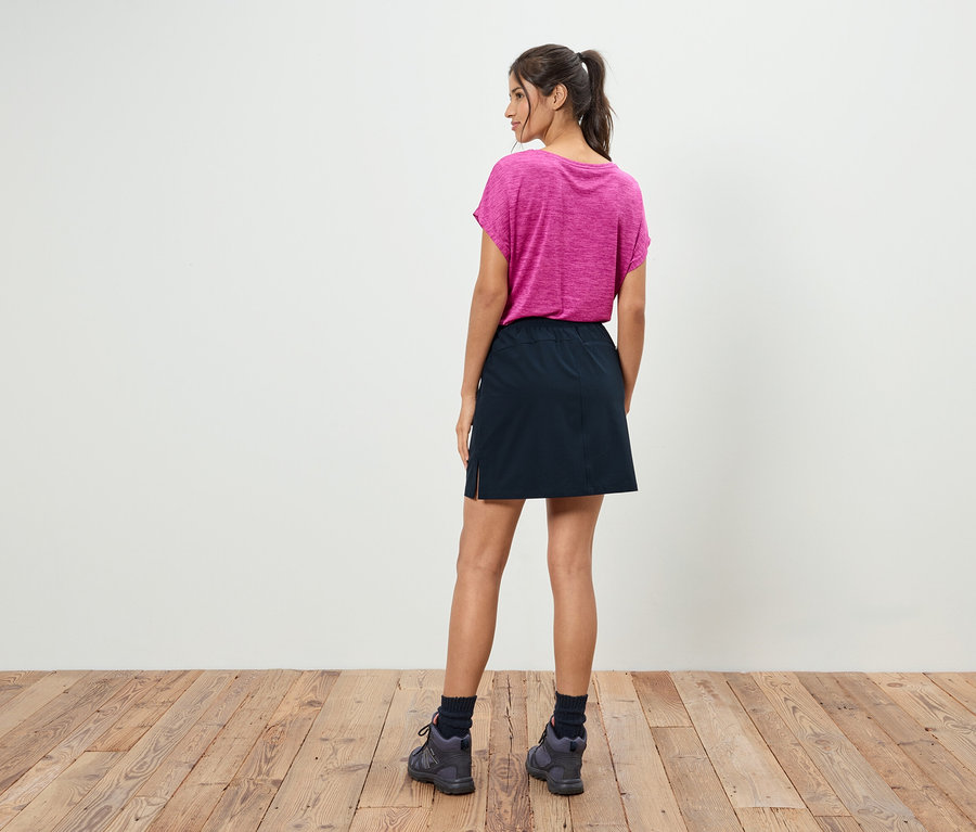 Žena s culíkem stojí zády k divákovi a má na sobě růžové tričko, černou sukni a šedé boty.