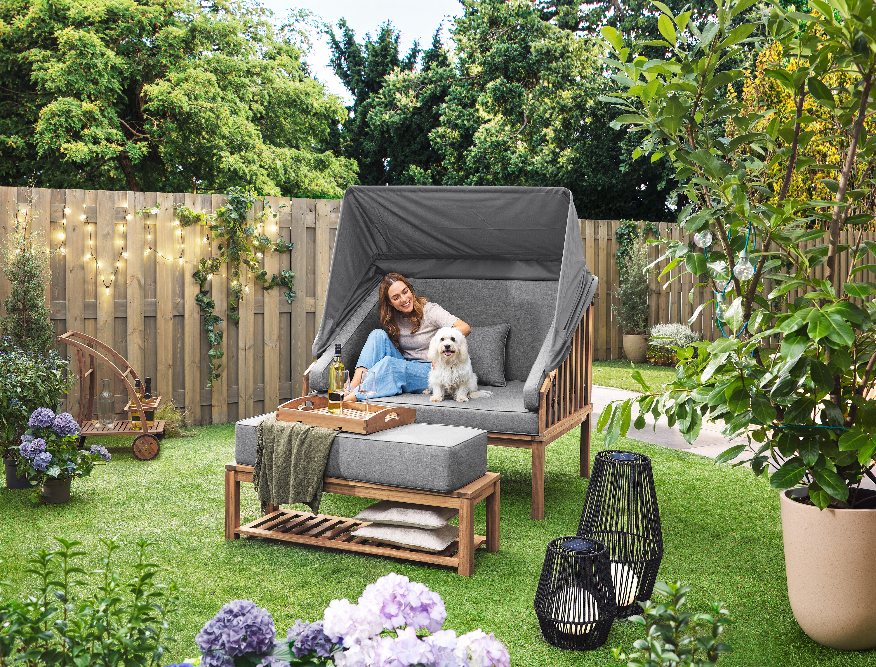 Žena sedí se psem v plážovém koši Premium Lounge-Strandkorb mit Sonnenschutz-Dach. Na trávníku stojí solární lucerna s textilním výpletem, velká a solární lucerna s textilním výpletem, malá. V pozadí je barový vozík s odnímatelným podnosem. Na plotě visí venkovní světelný řetěz s LED a síťovým adaptérem.