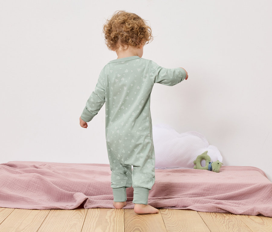 Malé dítě stojí zády ke kameře na posteli s růžovou dekou a zelenou hračkou.