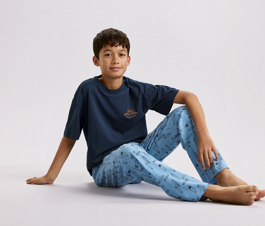 Chlapec sedí na zemi, má na sobě tmavě modré tričko s malým sluncem a vlnami a světle modré pyžamové kalhoty se vzorem surfových prken.
