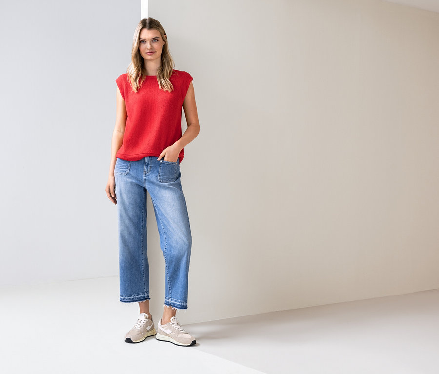 Žena stojí ve světlé místnosti, má na sobě červený top, modré džíny a tenisky.