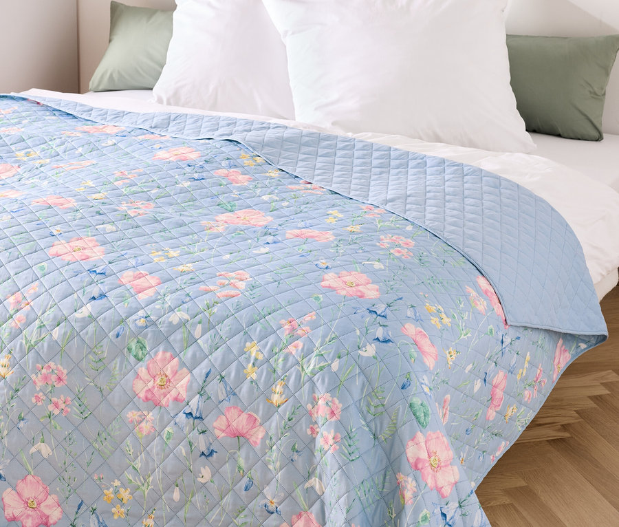 Modrý přehoz s květinovým vzorem na posteli s bílými polštáři.